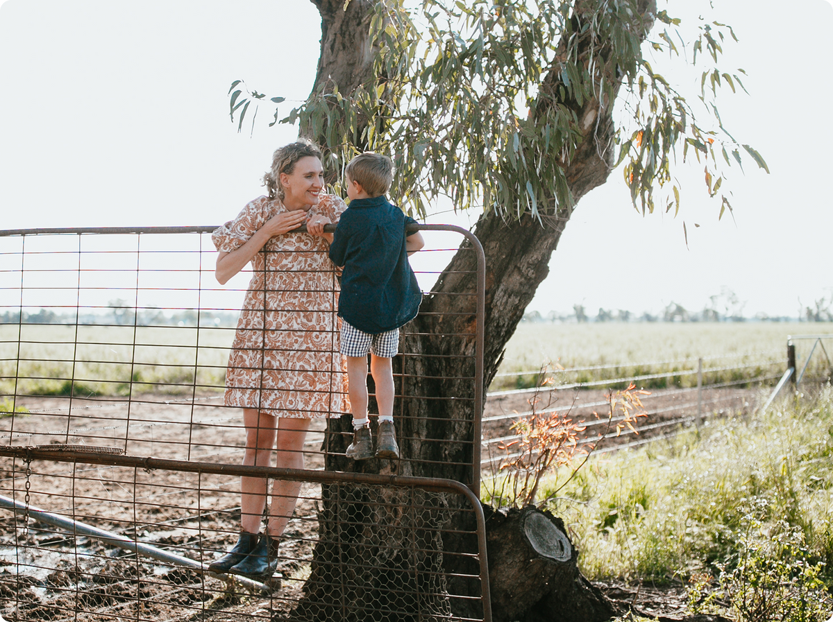 A woman in a patterned dress and a young boy in a blue shirt stand on a metal gate next to a large tree in an outdoor rural setting.