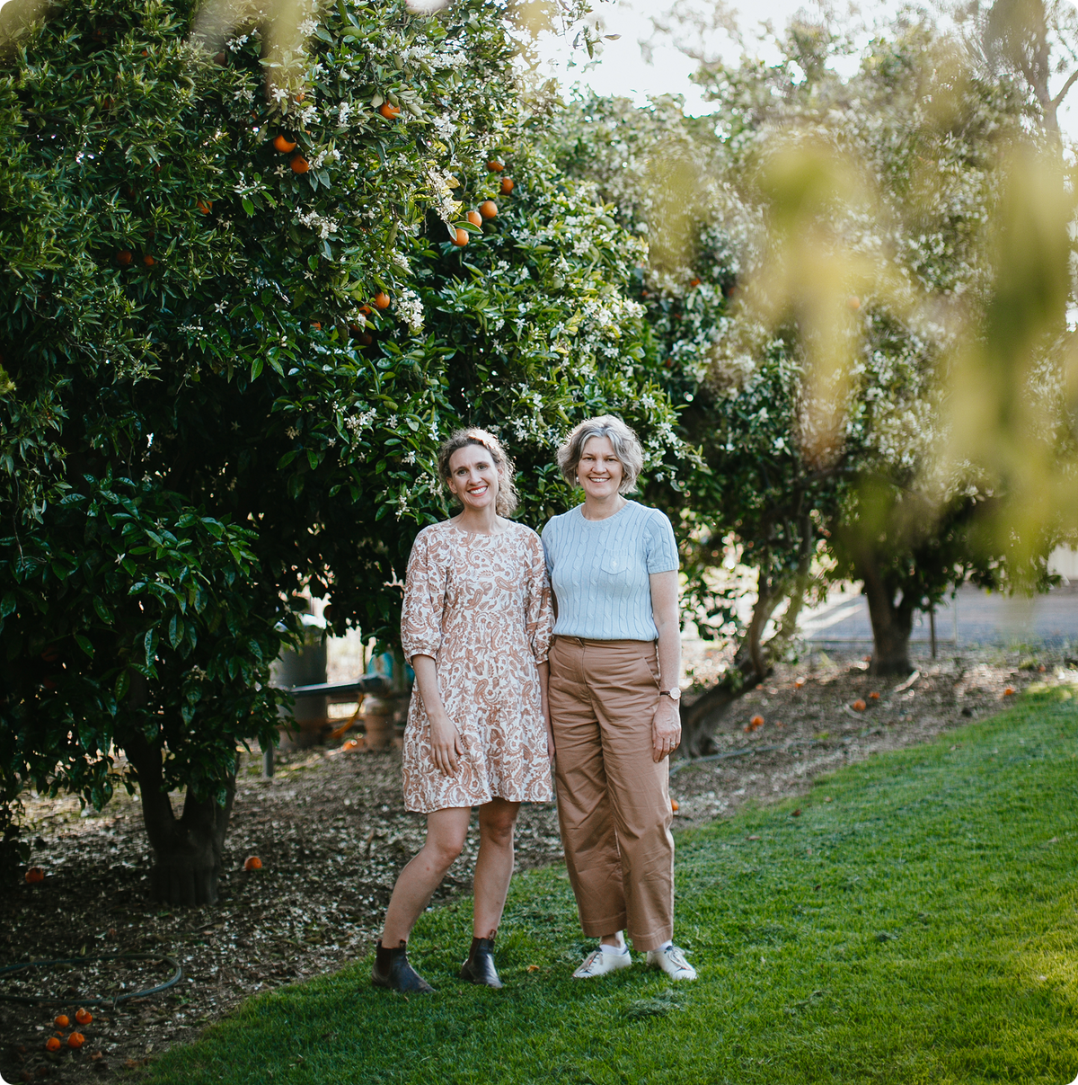 Two women standing outdoors in front of leafy trees with fruit, wearing casual dresses and tops in a grassy yard setting.