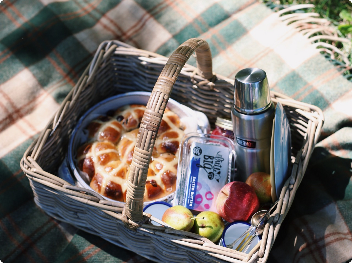 A wicker picnic basket filled with hot cross buns, a silver thermos, a container of fruit, and apples, set on a green and beige plaid blanket outdoors.